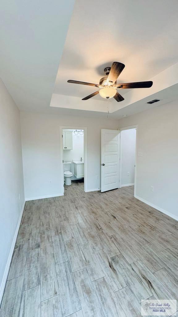 unfurnished bedroom featuring light wood-type flooring, ensuite bathroom, a tray ceiling, and ceiling fan