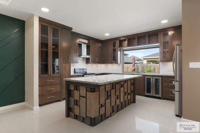 kitchen featuring wall chimney range hood, stainless steel appliances, dark brown cabinetry, a kitchen island, and beverage cooler