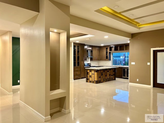 kitchen featuring wall chimney range hood, a center island, dark brown cabinetry, decorative backsplash, and a raised ceiling