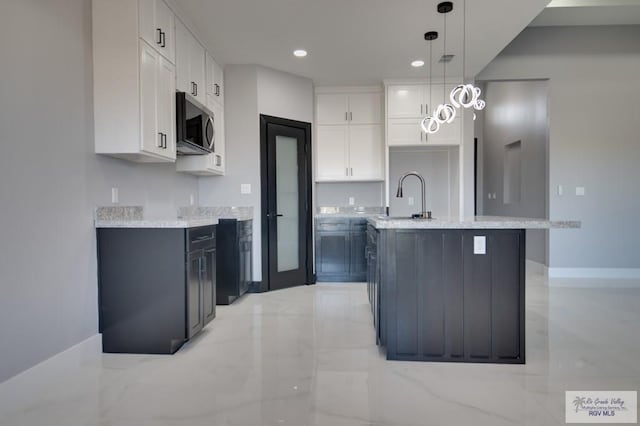 kitchen featuring stainless steel microwave, decorative light fixtures, a center island with sink, marble finish floor, and white cabinetry