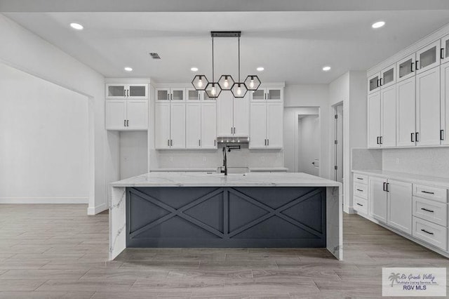 kitchen with white cabinetry, a center island with sink, hanging light fixtures, light stone countertops, and sink