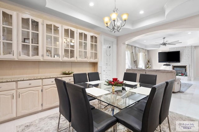 dining room with ceiling fan with notable chandelier, light tile patterned flooring, a raised ceiling, and a fireplace