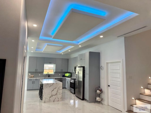 kitchen with gray cabinetry, a raised ceiling, decorative backsplash, appliances with stainless steel finishes, and a kitchen island