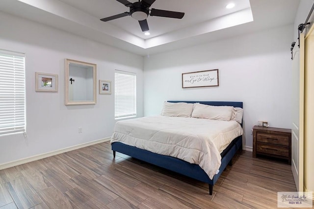 bedroom with ceiling fan, a barn door, wood-type flooring, and a tray ceiling