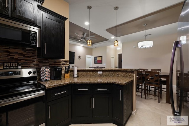 kitchen with hanging light fixtures, dark stone countertops, appliances with stainless steel finishes, a tray ceiling, and backsplash