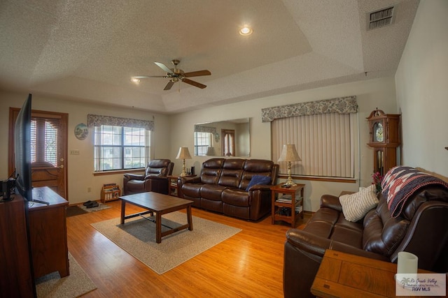 living room with a raised ceiling, light wood-type flooring, a textured ceiling, and ceiling fan