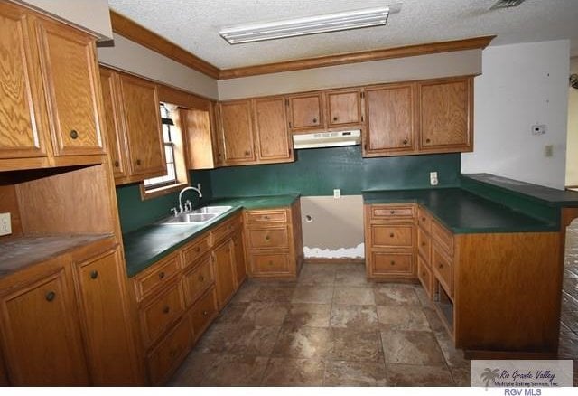 kitchen featuring brown cabinetry, a peninsula, a textured ceiling, under cabinet range hood, and a sink