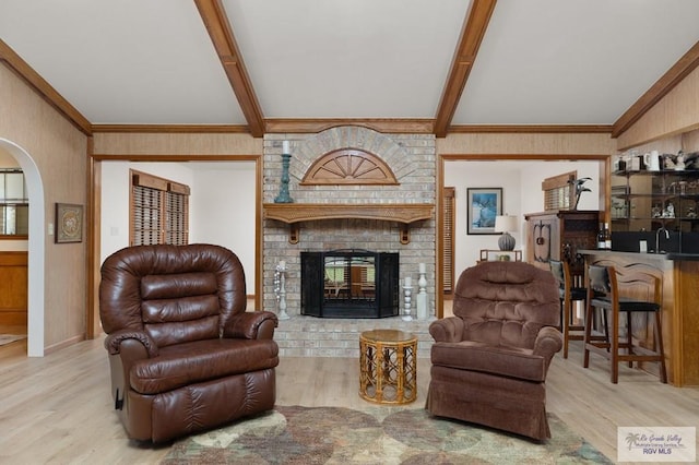 living room featuring arched walkways, a brick fireplace, crown molding, beamed ceiling, and light wood-style flooring