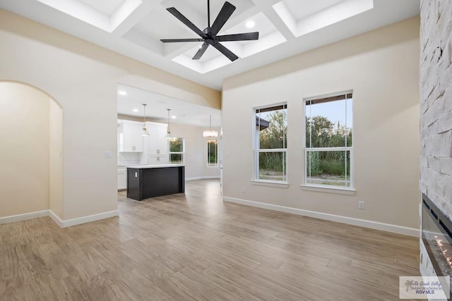 unfurnished living room featuring light wood-type flooring, coffered ceiling, ceiling fan, beam ceiling, and a stone fireplace