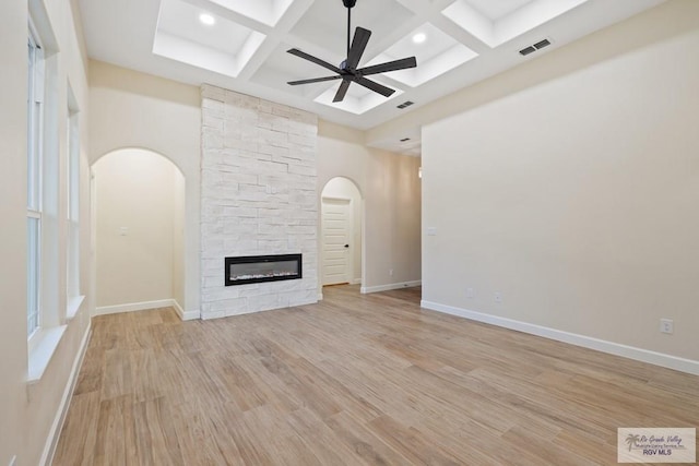 unfurnished living room featuring light wood-type flooring, coffered ceiling, ceiling fan, beamed ceiling, and a stone fireplace