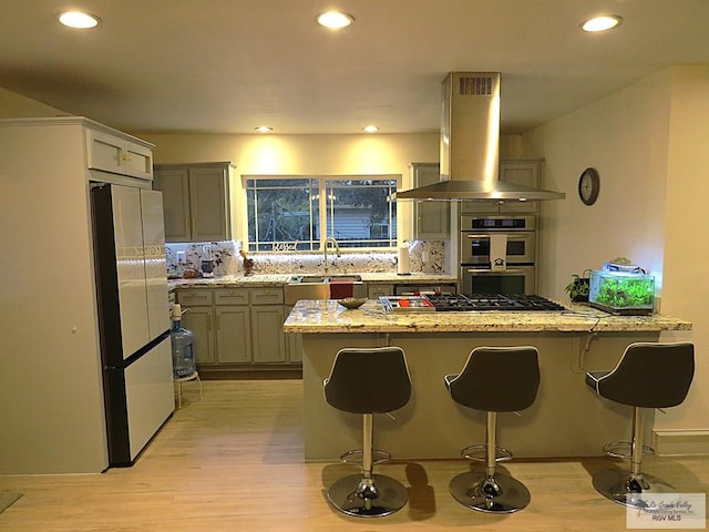 kitchen with light stone countertops, backsplash, island range hood, stainless steel appliances, and light hardwood / wood-style floors