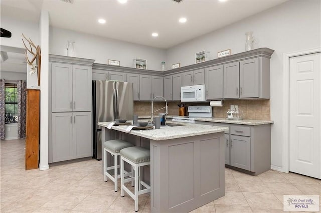 kitchen featuring white appliances, a center island with sink, gray cabinets, tasteful backsplash, and light stone counters