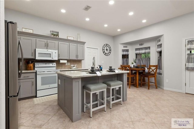 kitchen featuring light stone countertops, backsplash, gray cabinetry, white appliances, and a center island with sink