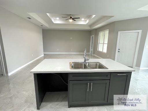 kitchen featuring gray cabinetry, a tray ceiling, ceiling fan, sink, and a center island with sink