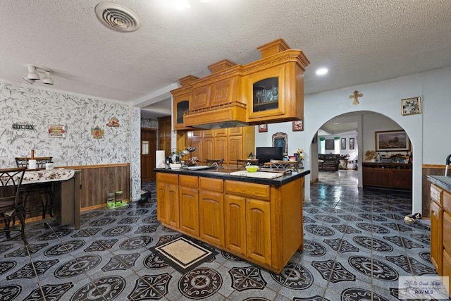 kitchen featuring glass insert cabinets, a textured ceiling, wallpapered walls, and wainscoting