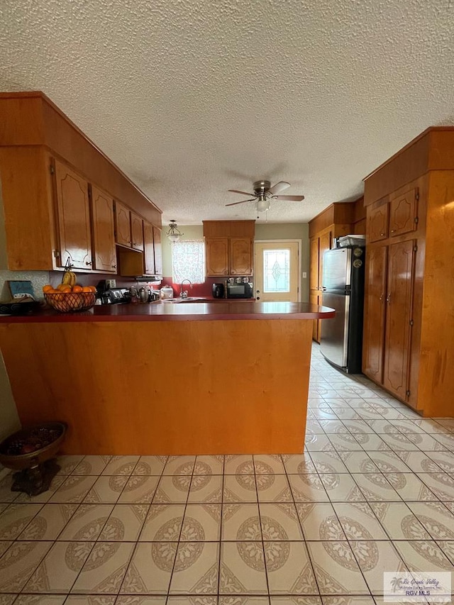 kitchen featuring brown cabinets, a sink, freestanding refrigerator, a peninsula, and ceiling fan