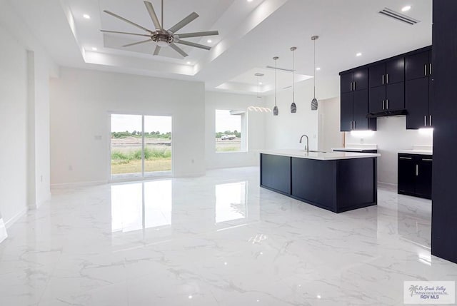 kitchen with a tray ceiling, hanging light fixtures, a center island with sink, and ceiling fan