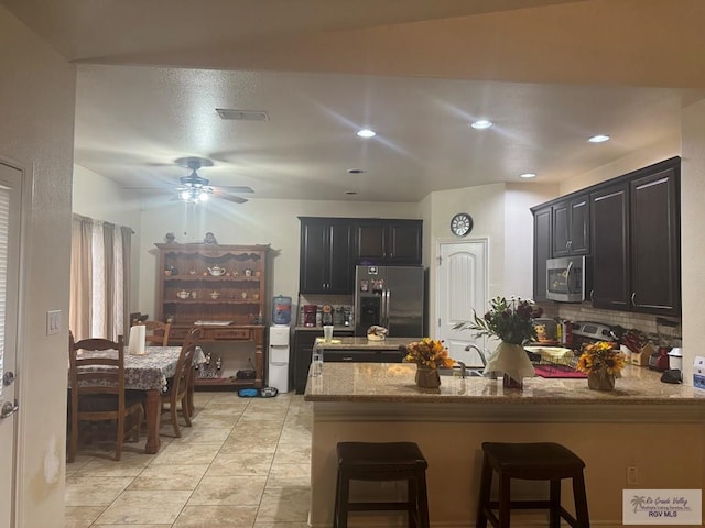 kitchen featuring a peninsula, light stone counters, a kitchen bar, a ceiling fan, and appliances with stainless steel finishes