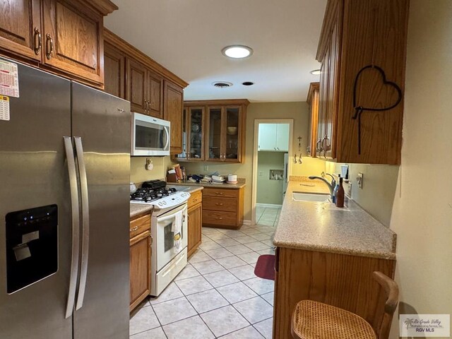 kitchen with stainless steel appliances, a sink, light tile patterned flooring, brown cabinets, and glass insert cabinets