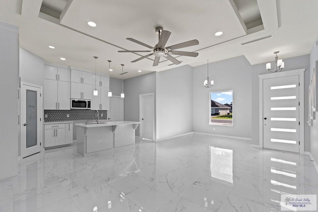 kitchen featuring ceiling fan with notable chandelier, pendant lighting, tasteful backsplash, white cabinets, and a kitchen island with sink