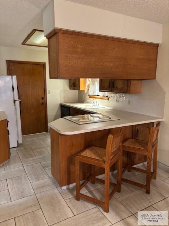 kitchen featuring a textured ceiling, a peninsula, light countertops, freestanding refrigerator, and brown cabinetry