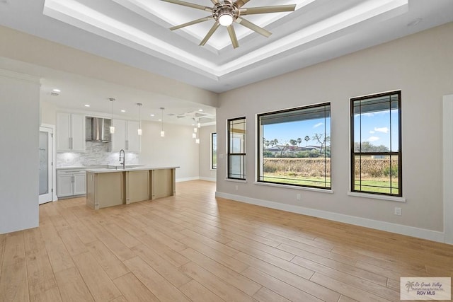 unfurnished living room with ceiling fan, sink, a tray ceiling, and light hardwood / wood-style floors