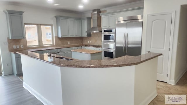 kitchen with wall chimney range hood, built in fridge, dark stone countertops, decorative backsplash, and a kitchen island