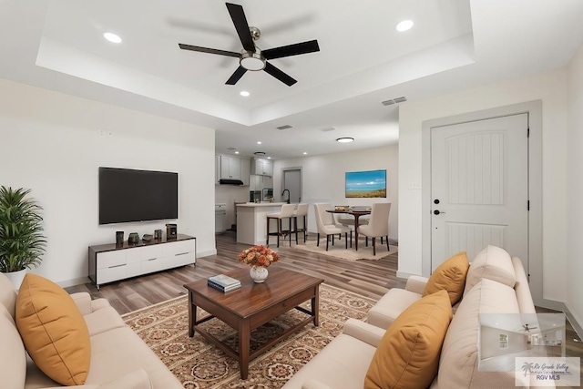 living room with ceiling fan, sink, a tray ceiling, and hardwood / wood-style floors