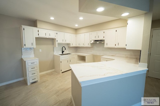 kitchen with a sink, a peninsula, under cabinet range hood, white cabinetry, and backsplash