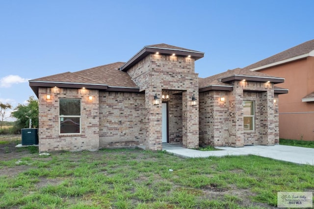 prairie-style home featuring central air condition unit, brick siding, roof with shingles, and a front yard