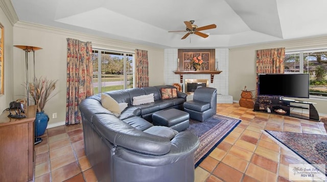 tiled living area with a ceiling fan, baseboards, ornamental molding, a raised ceiling, and a brick fireplace