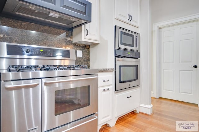 kitchen with appliances with stainless steel finishes, dark stone counters, ventilation hood, white cabinets, and light hardwood / wood-style floors