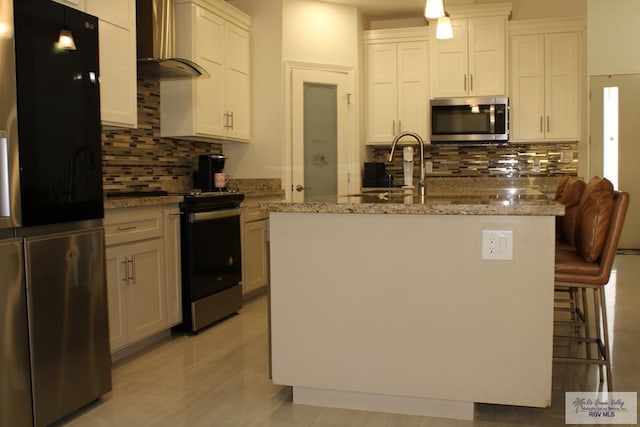 kitchen with backsplash, wall chimney range hood, light stone countertops, an island with sink, and stainless steel appliances