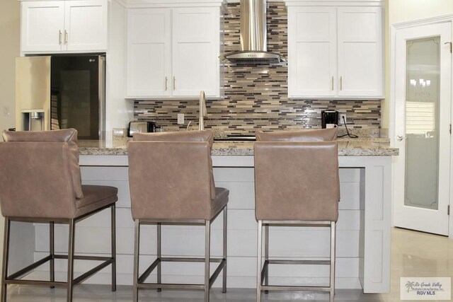 3512 Westcreek Dr, Brownsville, TX 78526 - kitchen featuring wall chimney range hood, fridge with ice dispenser, decorative backsplash, white cabinets, and a kitchen bar