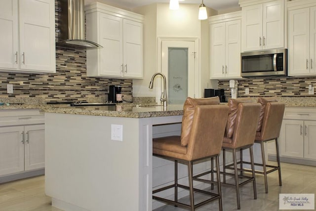 3512 Westcreek Dr, Brownsville, TX 78526 - kitchen featuring stainless steel microwave, wall chimney range hood, tasteful backsplash, a breakfast bar, and light stone countertops