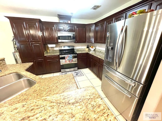 kitchen with appliances with stainless steel finishes, a sink, light tile patterned floors, dark brown cabinetry, and light stone countertops