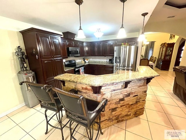 kitchen featuring stainless steel appliances, arched walkways, light tile patterned floors, dark brown cabinetry, and a breakfast bar