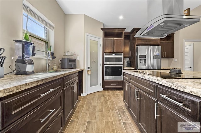 kitchen with island exhaust hood, appliances with stainless steel finishes, light stone counters, dark brown cabinetry, and sink