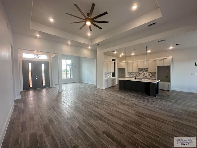 unfurnished living room with dark hardwood / wood-style floors, sink, ceiling fan with notable chandelier, and a tray ceiling