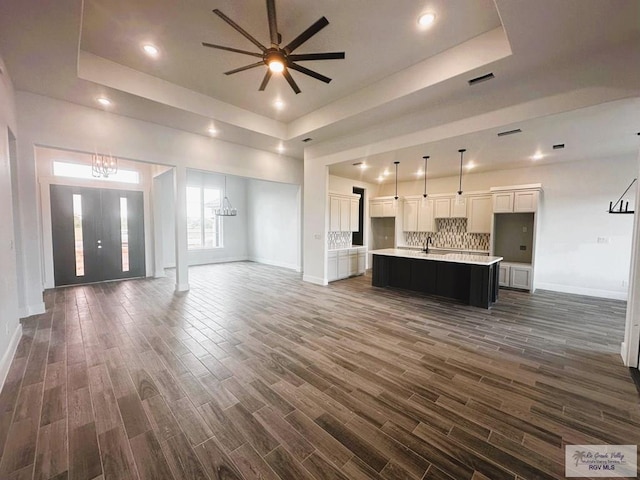 unfurnished living room with ceiling fan with notable chandelier, sink, dark wood-type flooring, and a tray ceiling