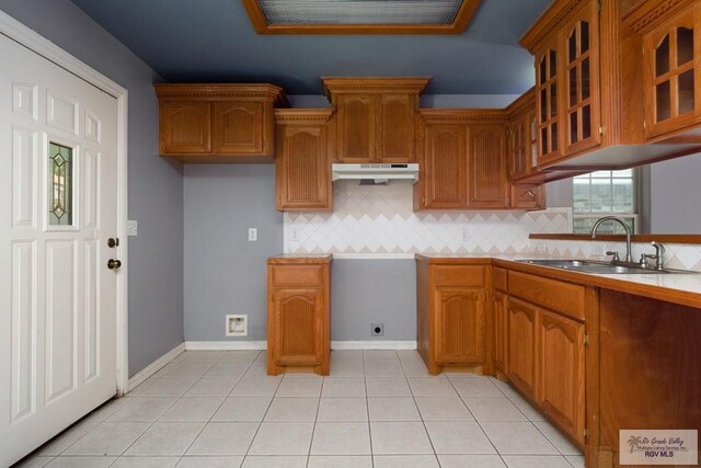kitchen featuring backsplash, brown cabinets, a sink, glass insert cabinets, and under cabinet range hood