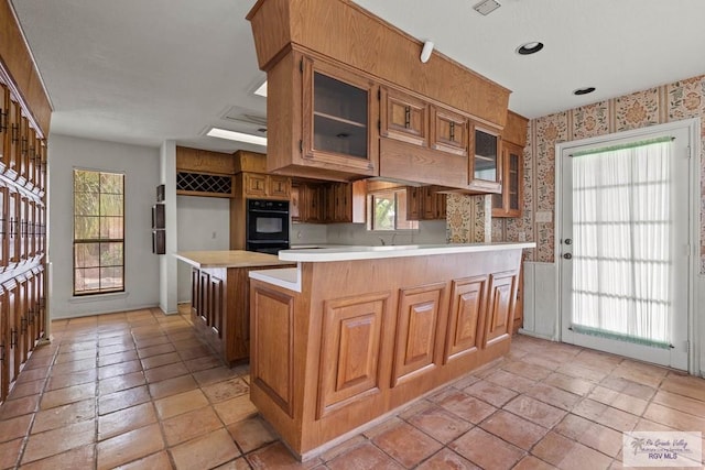 kitchen featuring light countertops, brown cabinetry, a peninsula, wallpapered walls, and glass insert cabinets