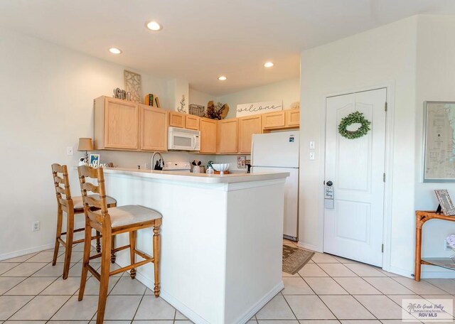 kitchen with light tile patterned floors, white appliances, kitchen peninsula, and light brown cabinetry