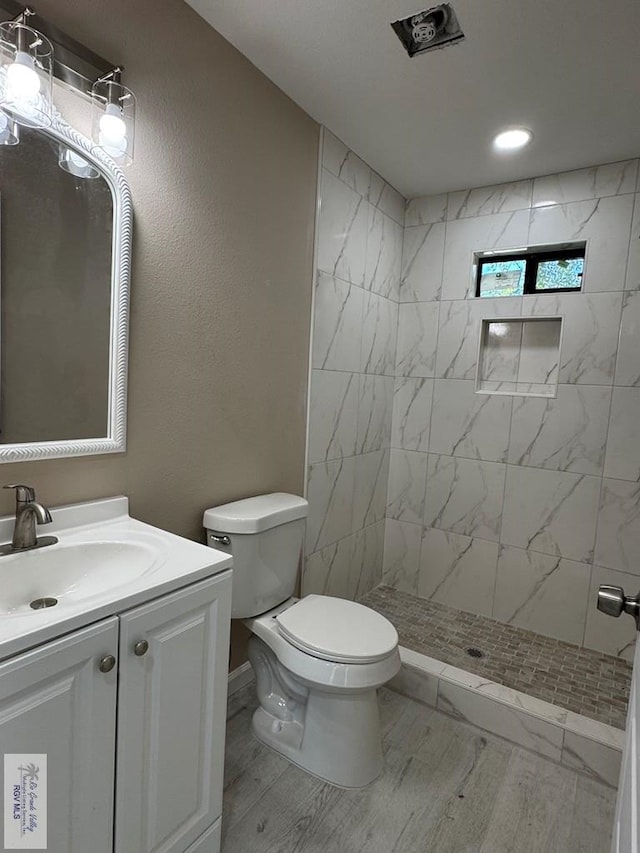 bathroom with wood finished floors, toilet, vanity, a tile shower, and recessed lighting