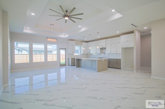 kitchen featuring a raised ceiling, a kitchen island with sink, sink, white cabinets, and hanging light fixtures