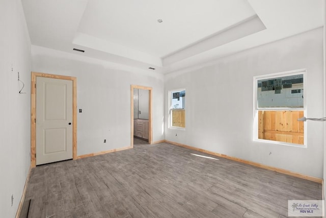 unfurnished bedroom with a tray ceiling, visible vents, ensuite bathroom, wood finished floors, and baseboards