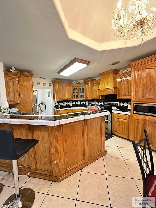 kitchen featuring tile countertops, appliances with stainless steel finishes, brown cabinetry, and custom range hood