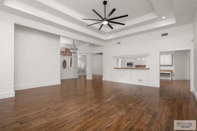 unfurnished living room with decorative columns, ceiling fan with notable chandelier, a raised ceiling, dark wood-type flooring, and a high ceiling