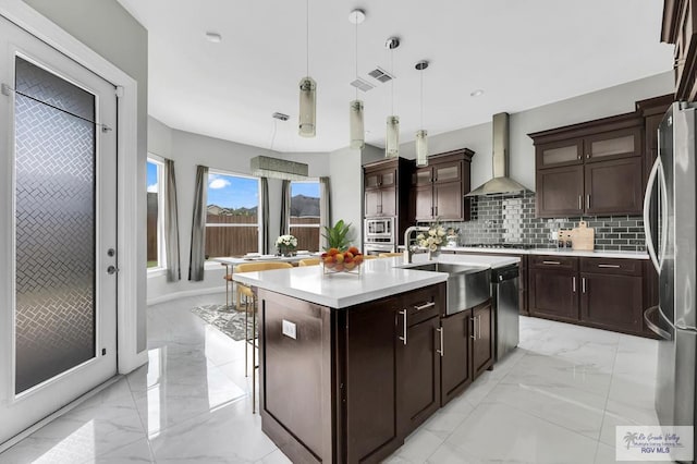 kitchen featuring appliances with stainless steel finishes, tasteful backsplash, wall chimney exhaust hood, pendant lighting, and an island with sink