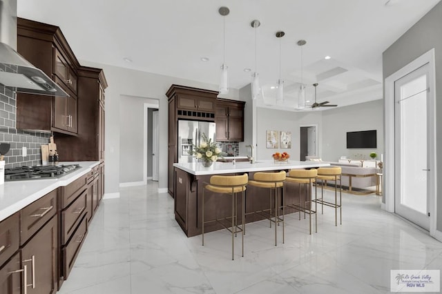 kitchen featuring a kitchen island with sink, hanging light fixtures, wall chimney exhaust hood, decorative backsplash, and stainless steel appliances